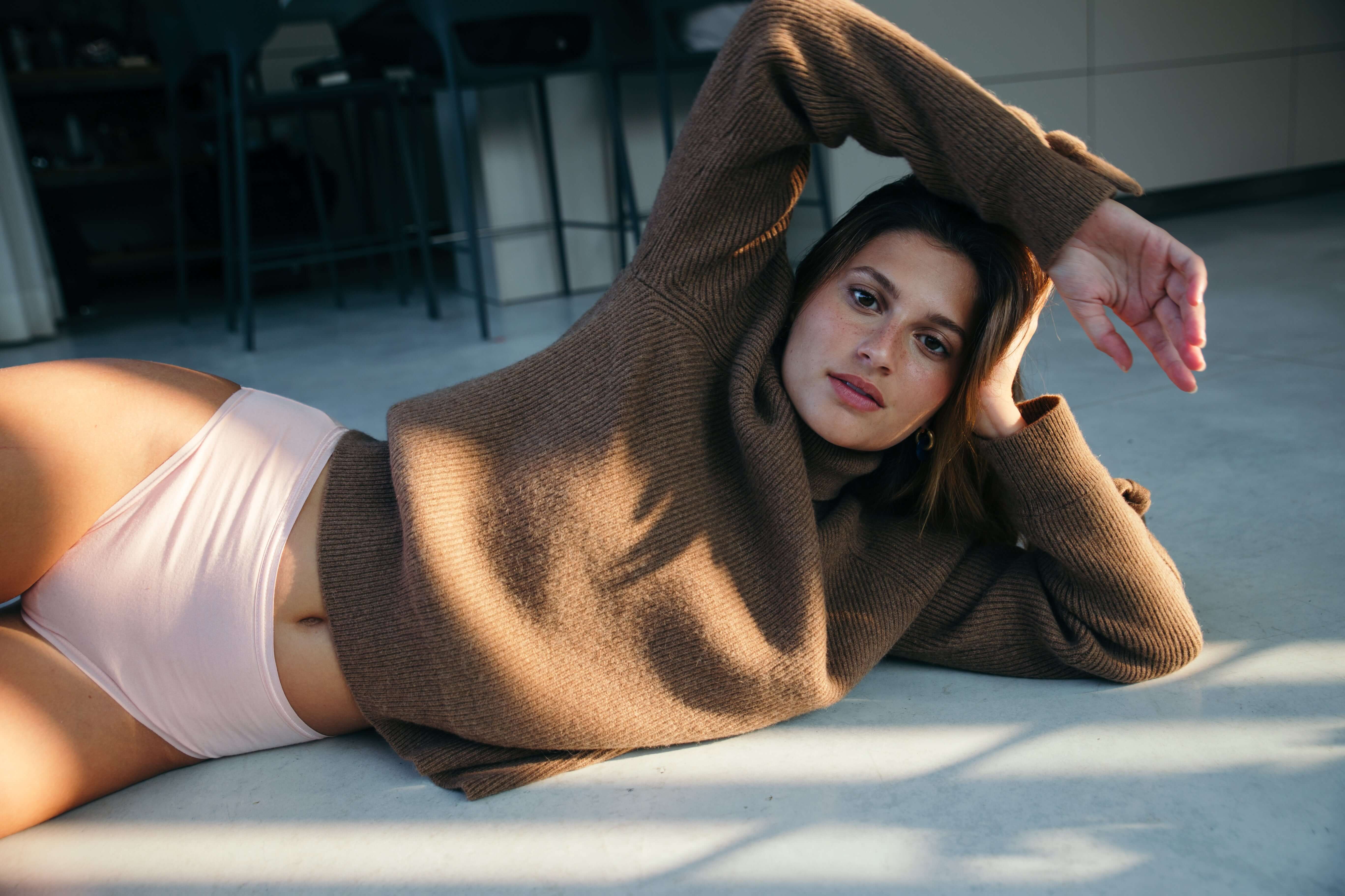 אישה לובשת תחתוני כותנה אורגנית שוכבת על רצפת חדר עם סוודר חום, מדגימה נוחות ובריאות בלבוש אינטימי טבעי.